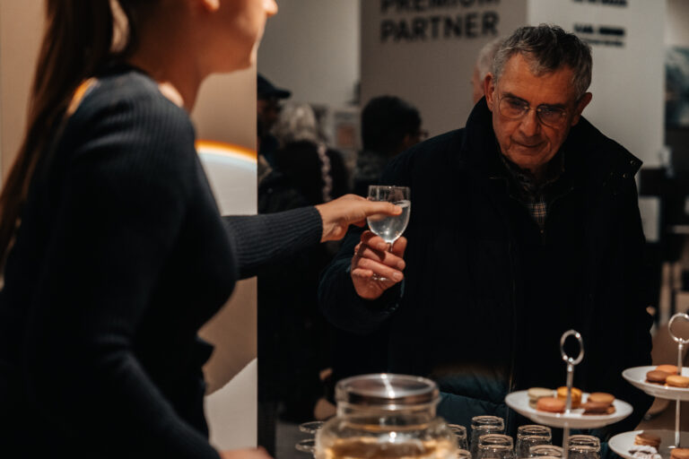 Een bezoeker wordt ontvangen met een drankje tijdens een demoavond